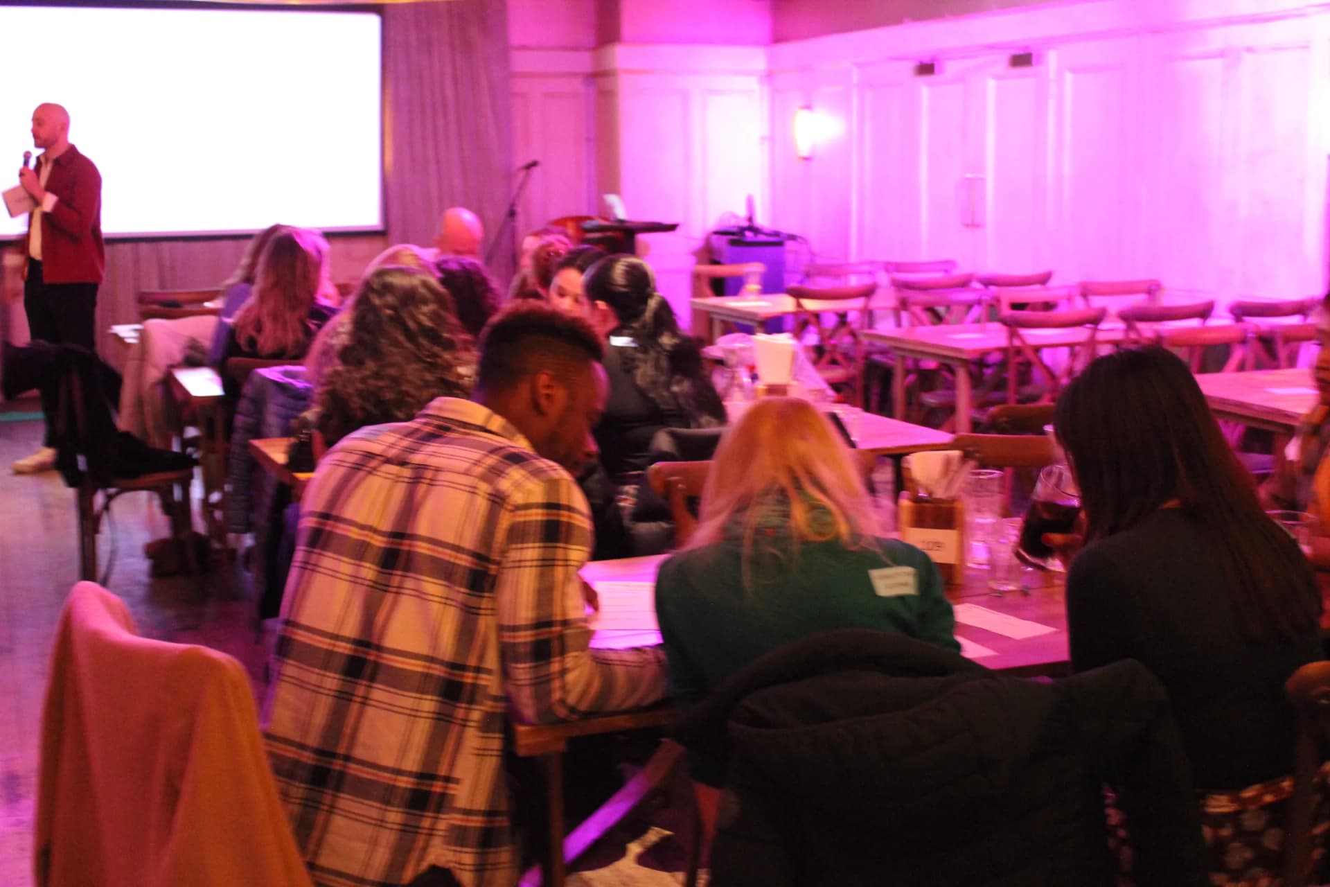 Host reading a question at the screen while teams write in the foreground.